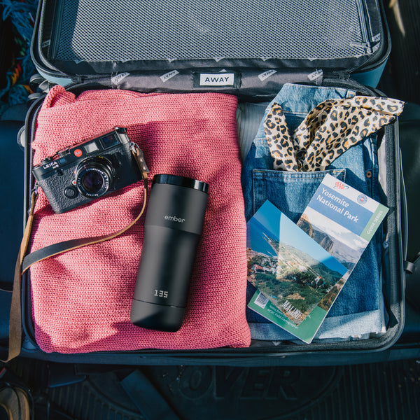 Suitcase filled with shirts, jeans, scarf, travel brochures, camera, and Ember Travel Mug.