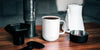 A white Ember Mug sits on a dark wooden table next to a white electric kettle and a black Aeropress with it's accessories. 