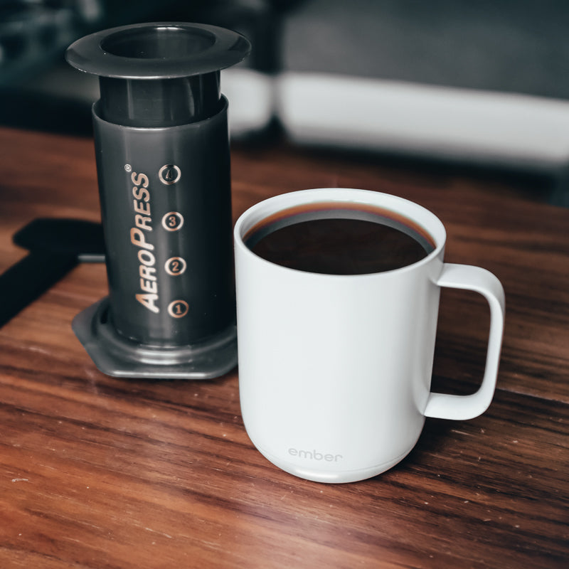 A white Ember Mug on a dark wood table with an AeroPress on the left.