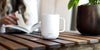 Tea brewing in an Ember Mug² in white on a dark wooden table with a woman reading a book in the background.