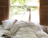 Person in bed under fluffy covers, next to a bright window, holding a white Ember Mug in the air.