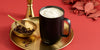 A Black Ember Mug² filled with heavy cream sits on a gold plate alongside a gold bowl filled with coffee beans and a small gold spoon. The red background contains a gold jigger and red leaves.