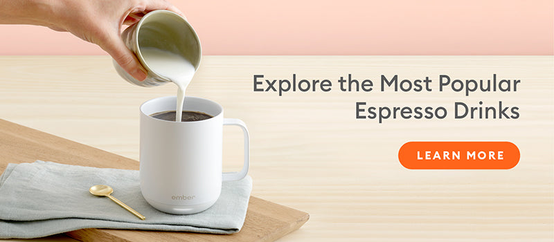 A hand pours cream from a small cup into a white Ember Mug² that's sitting on a blue cloth which is neatly folded and resting on a wooden cutting board. Next to the mug is a gold spoon, and there is a branch of green eucalyptus leaves in the background.