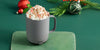 A Gray Ember Mug² is filled with whipped cream and topped with crushed candy cane and cinnamon. The mug sits on a dark green background filled with red and green holiday ornaments alongside a leafy branch with red berries.