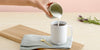 A hand pours cream from a small cup into a white Ember Mug² that's sitting on a blue cloth which is neatly folded and resting on a wooden cutting board. Next to the mug is a gold spoon, and there is a branch of green eucalyptus leaves in the background.